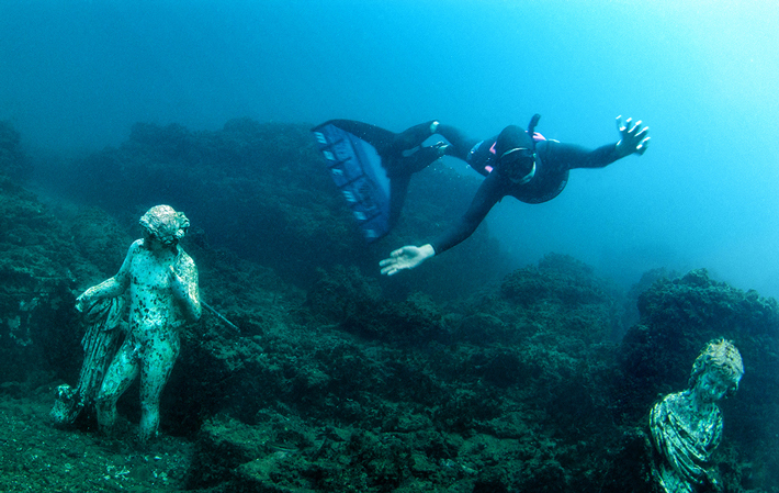 A diver swims between replicas of statues depicting Dionysus (left) and a young girl (right) at Baiae. (Pasquale Vassallo)